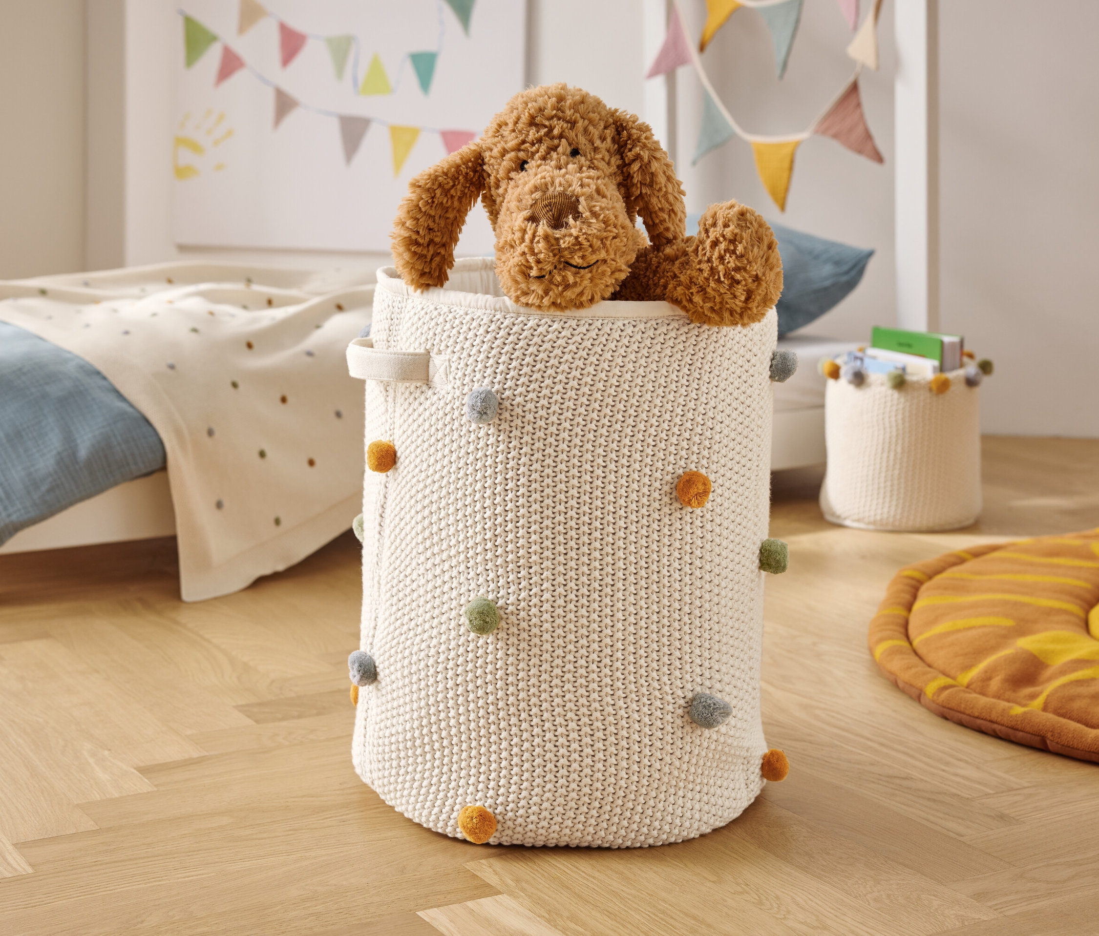 Brązowy pluszowy pies siedzi w białym, dzierganym koszu z kolorowymi pomponami.