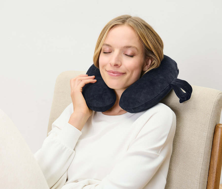 Kobieta siedzi z zamkniętymi oczami na krześle i ma na sobie ciemnoniebieską poduszkę podróżną.