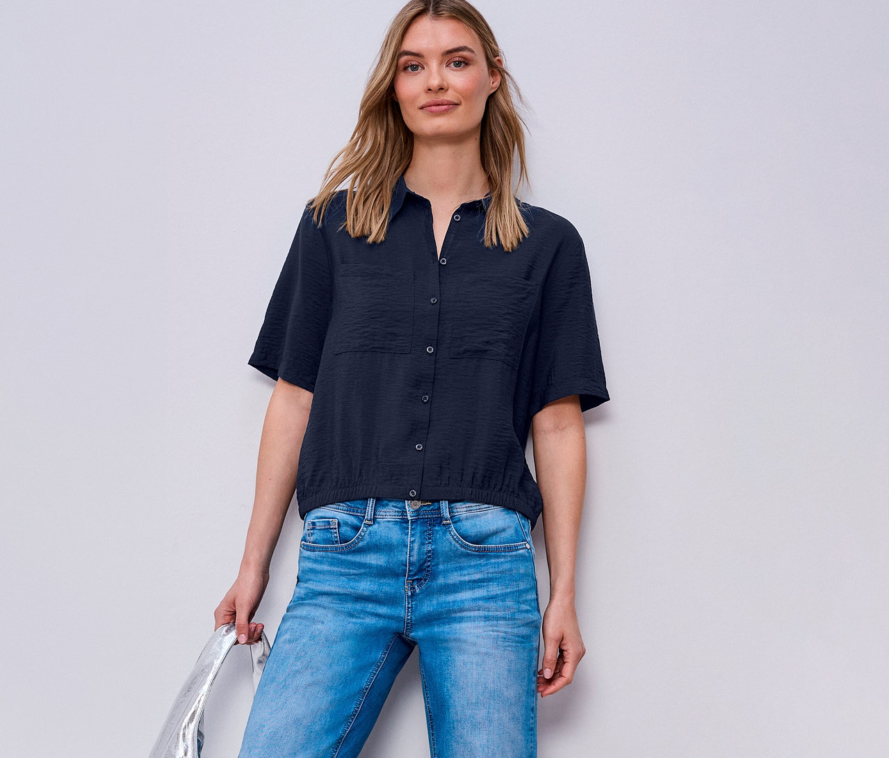 Kobieta ma na sobie dżinsy i koszulę Street One. Trzyma srebrną torbę.