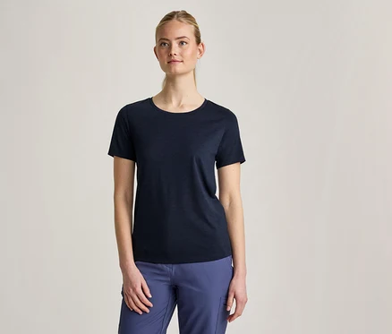 Kobieta z blond włosami spiętymi do tyłu, ubrana w granatowy T-shirt z krótkim rękawem i niebieskie spodnie, stoi na jasnoszarym tle.