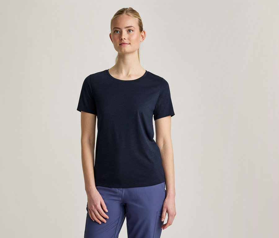 Kobieta z blond włosami spiętymi do tyłu, ubrana w granatowy T-shirt z krótkim rękawem i niebieskie spodnie, stoi na jasnoszarym tle.