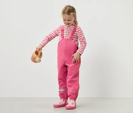 Dziewczynka podlewa wodę konewką, ubrana w różowe ogrodniczki i buty.
