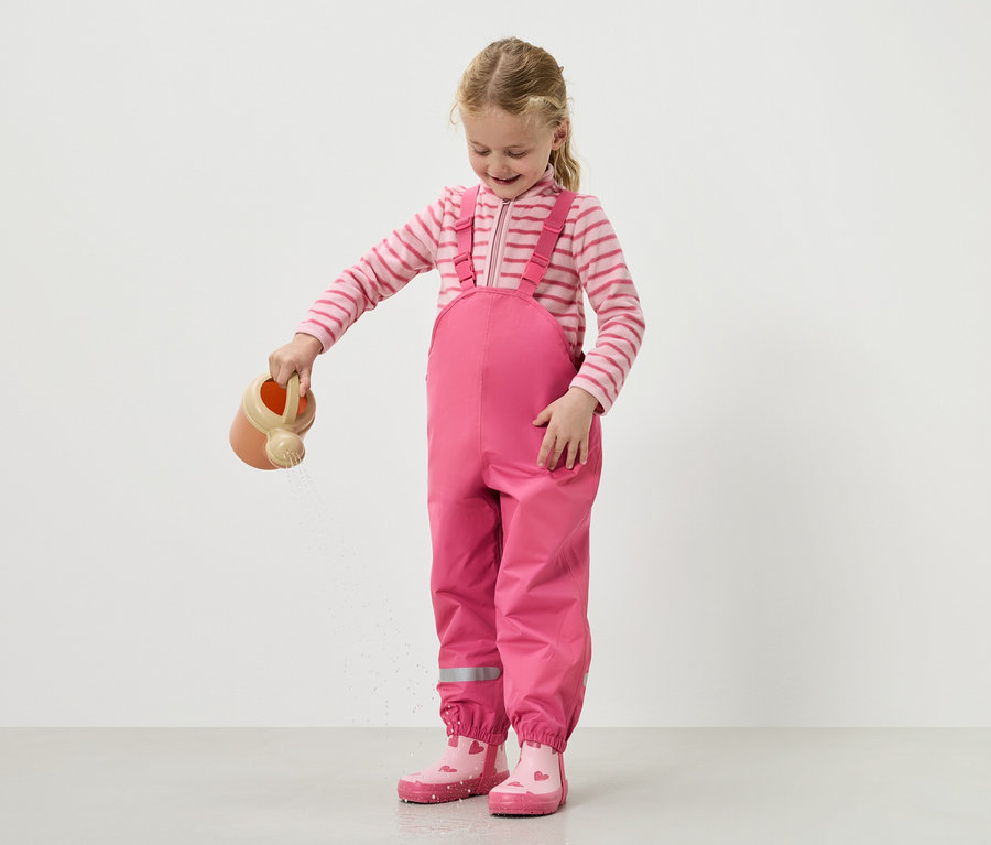 Dziewczynka podlewa wodę konewką, ubrana w różowe ogrodniczki i buty.