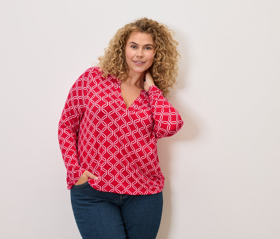 Kobieta o kręconych włosach w czerwono-białej bluzce i dżinsach, pozująca z ręką w kieszeni.