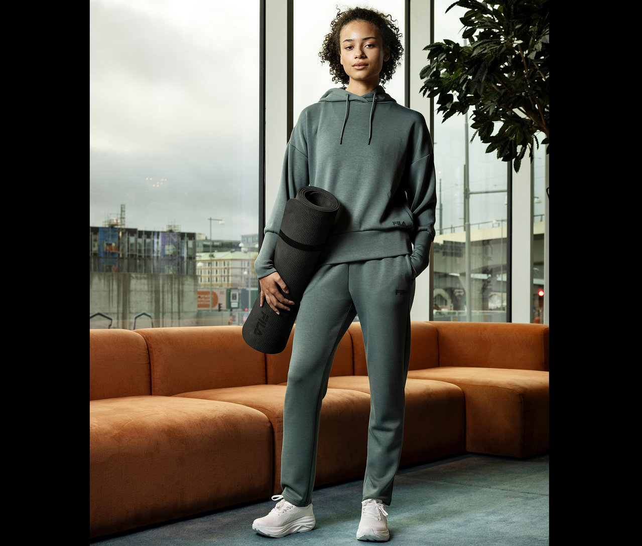 Młoda kobieta w szarym stroju sportowym trzyma zwiniętą matę do jogi na tle okna.