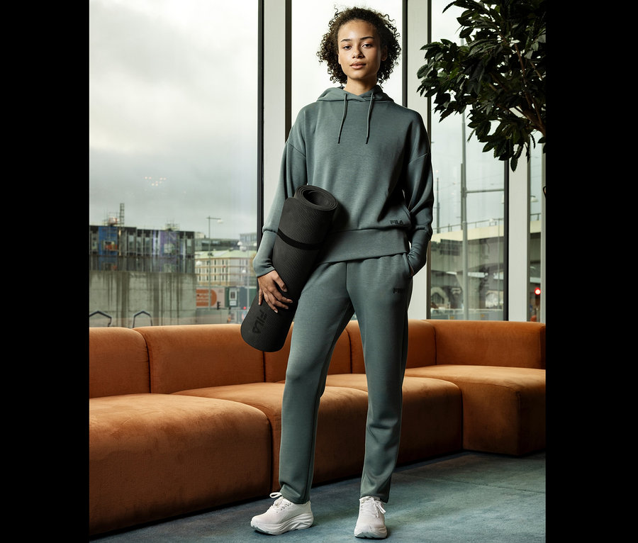 Młoda kobieta w szarym stroju sportowym trzyma zwiniętą matę do jogi na tle okna.