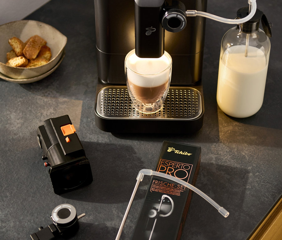 Ekspres »Esperto 2 Milk« Granite Black robi latte macchiato. Obok stoi regulator obrotowy pianki mlecznej, miska z pieczywem i opakowanie zestawu akcesoriów.