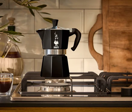 Czarny ekspres do kawy stoi na kuchence gazowej obok szklanki espresso.