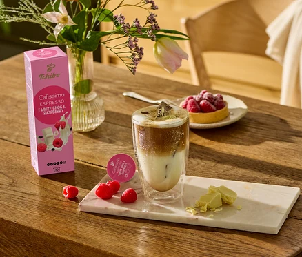 Na drewnianym stole stoi szklanka kawy, obok leżą kawałki białej czekolady i maliny na marmurowej desce. W tle widać opakowanie Cafissimo Flavoured Espresso – White Choc & Raspberry – 10 kapsułek.