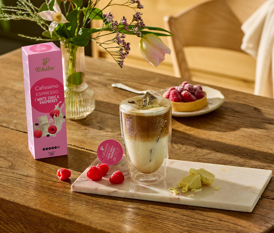Na drewnianym stole stoi szklanka kawy, obok leżą kawałki białej czekolady i maliny na marmurowej desce. W tle widać opakowanie Cafissimo Flavoured Espresso – White Choc & Raspberry – 10 kapsułek.