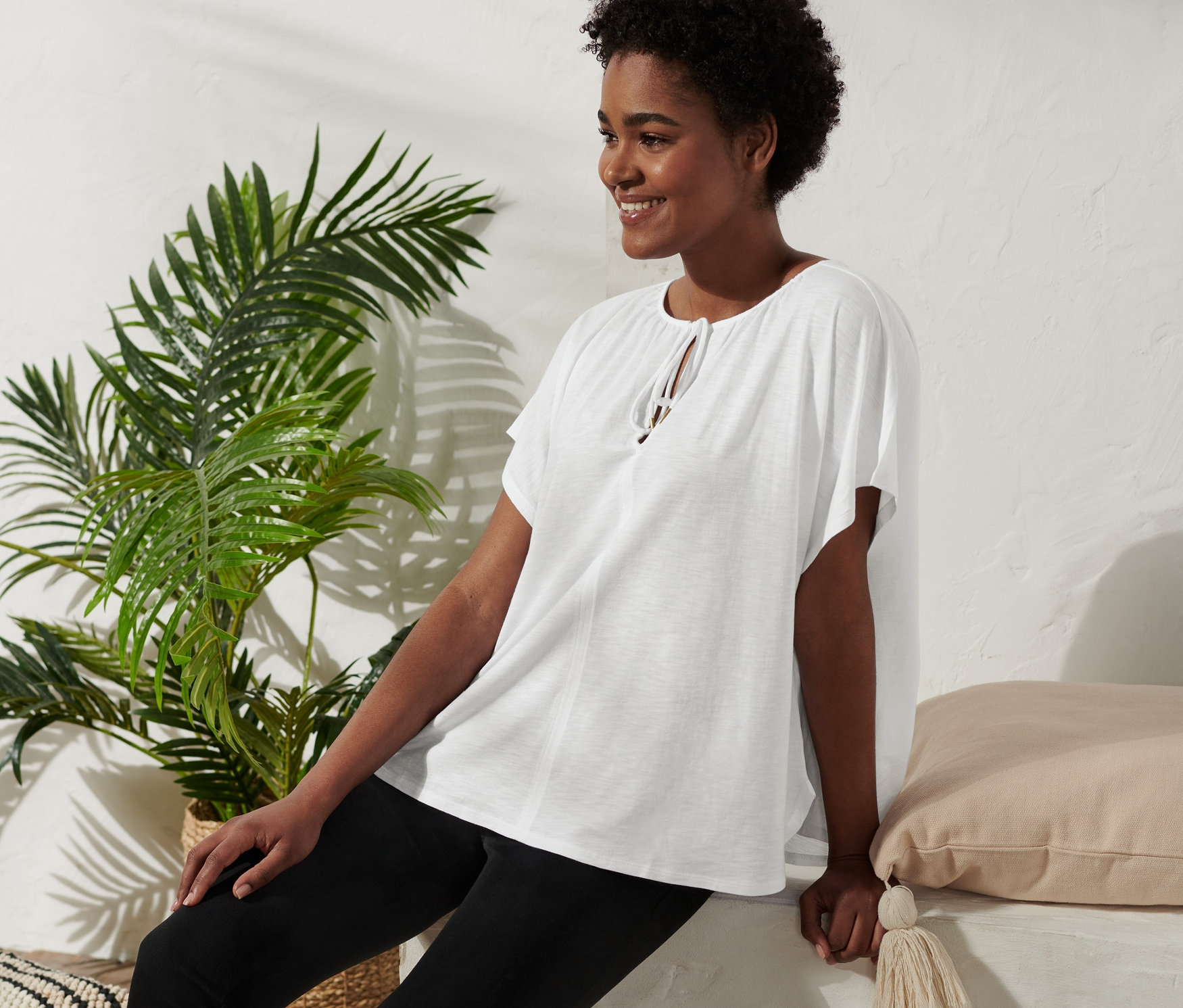 Kobieta siedzi i uśmiecha się, ma na sobie bluzkę z wiązaniem, czarne spodnie i sandały na platformie.