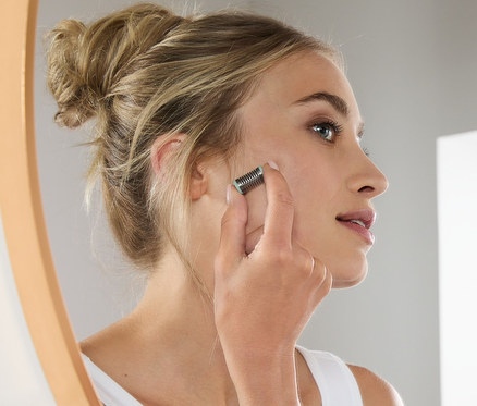 Kobieta używa mini depilatora do twarzy przed lustrem.