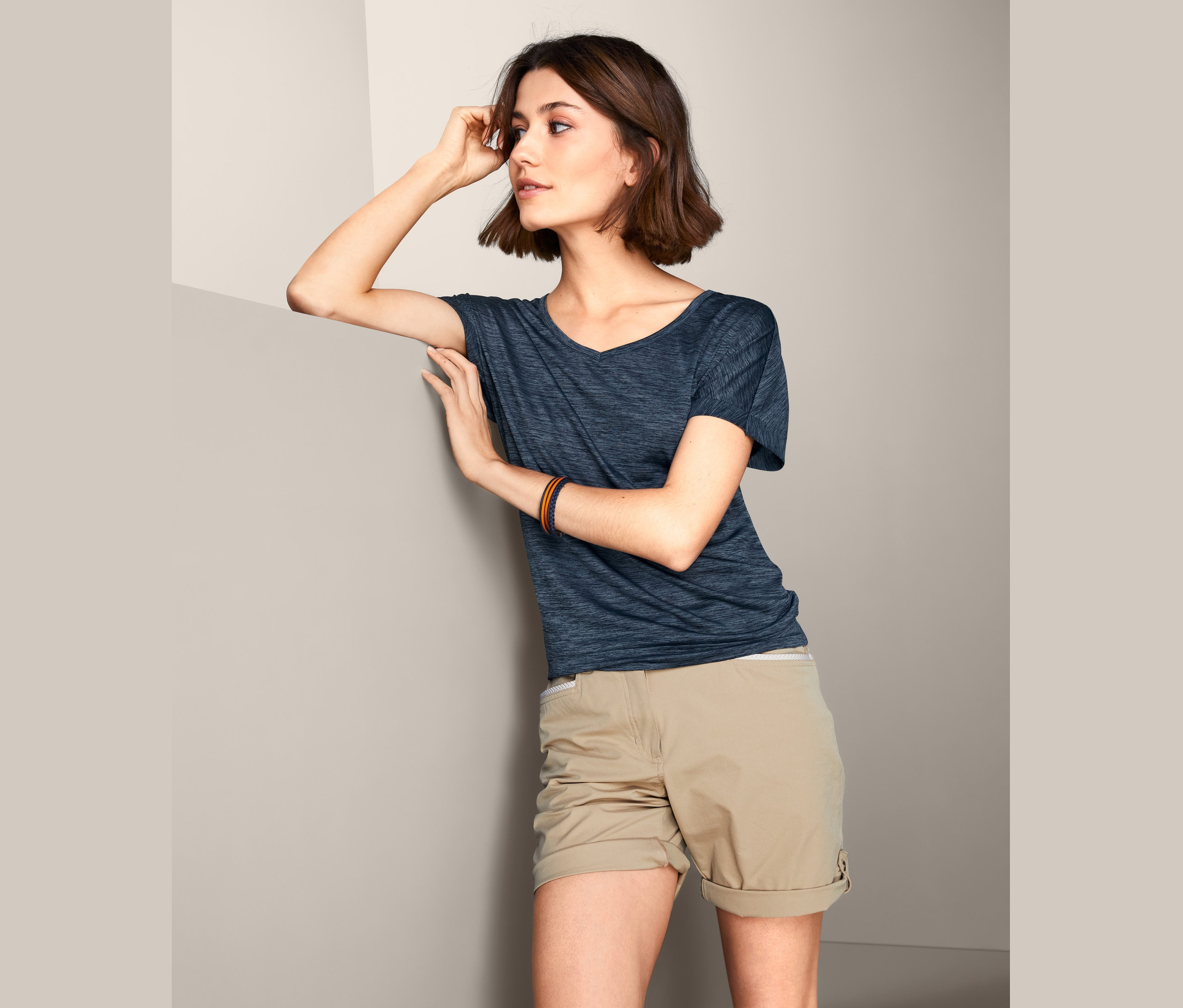 Kobieta oparta o ścianę ma na sobie granatową melanżową koszulkę funkcyjną i beżowe szorty.