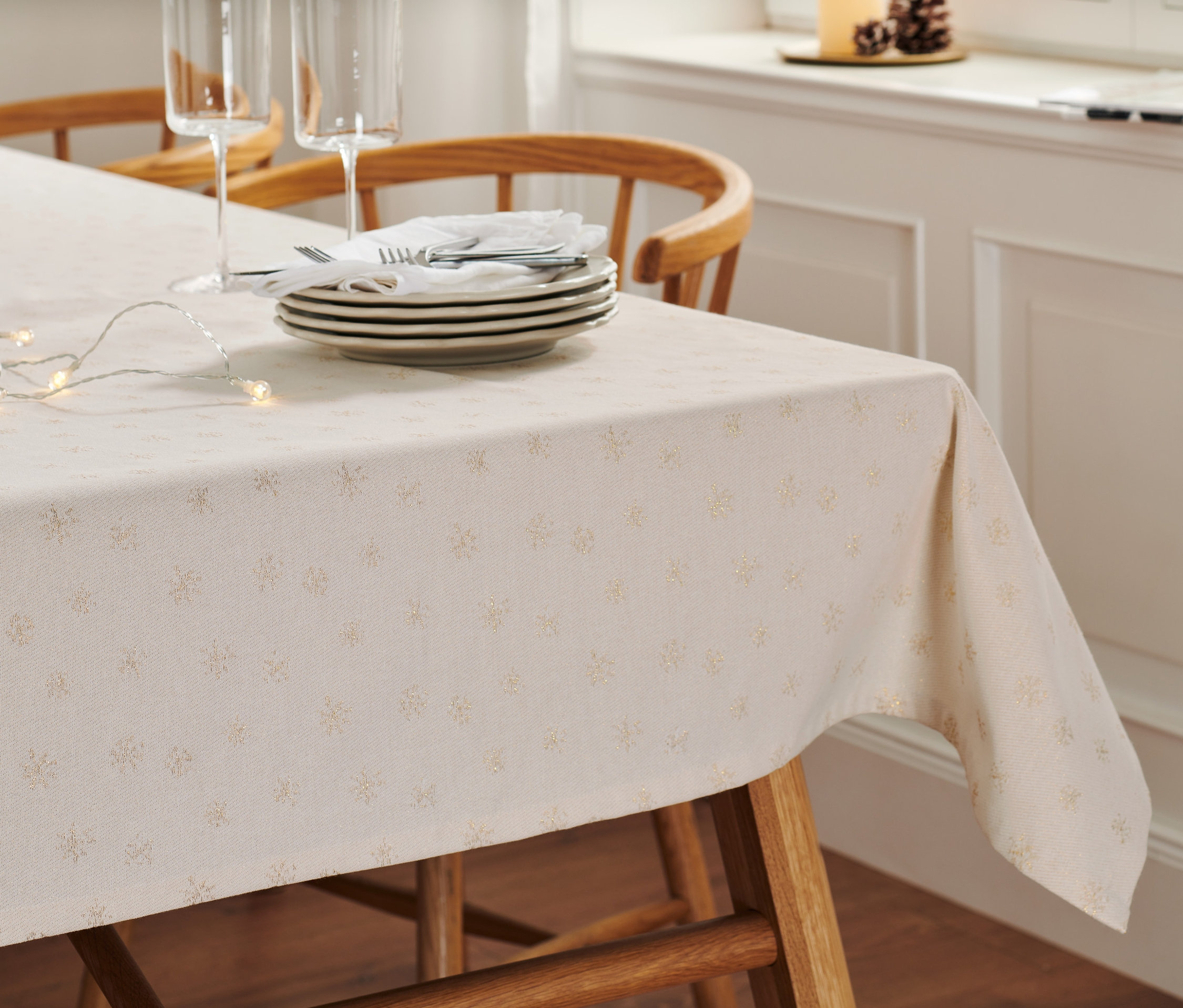 Na drewnianym stole leży obrus żakardowy premium z talerzami, sztućcami, szklankami i łańcuchem świetlnym.