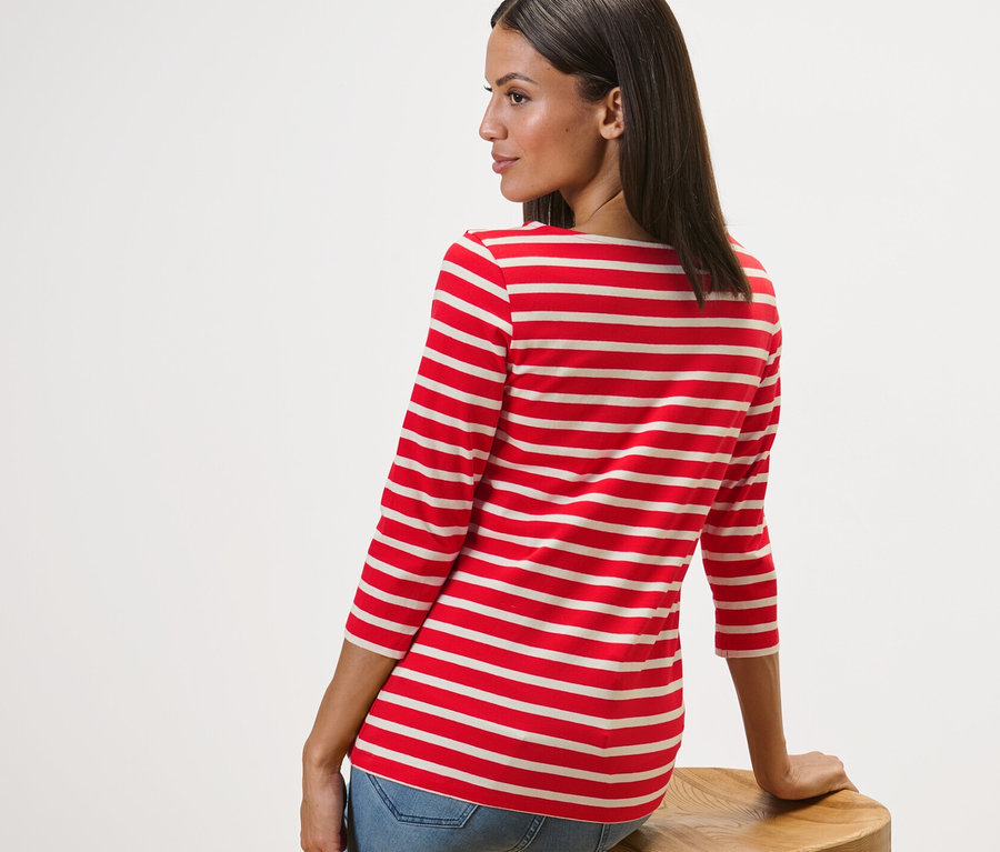 Kobieta siedzi bokiem na drewnianym stołku i ma na sobie bluzkę w paski z rękawami o długości 3/4, czerwone paski.