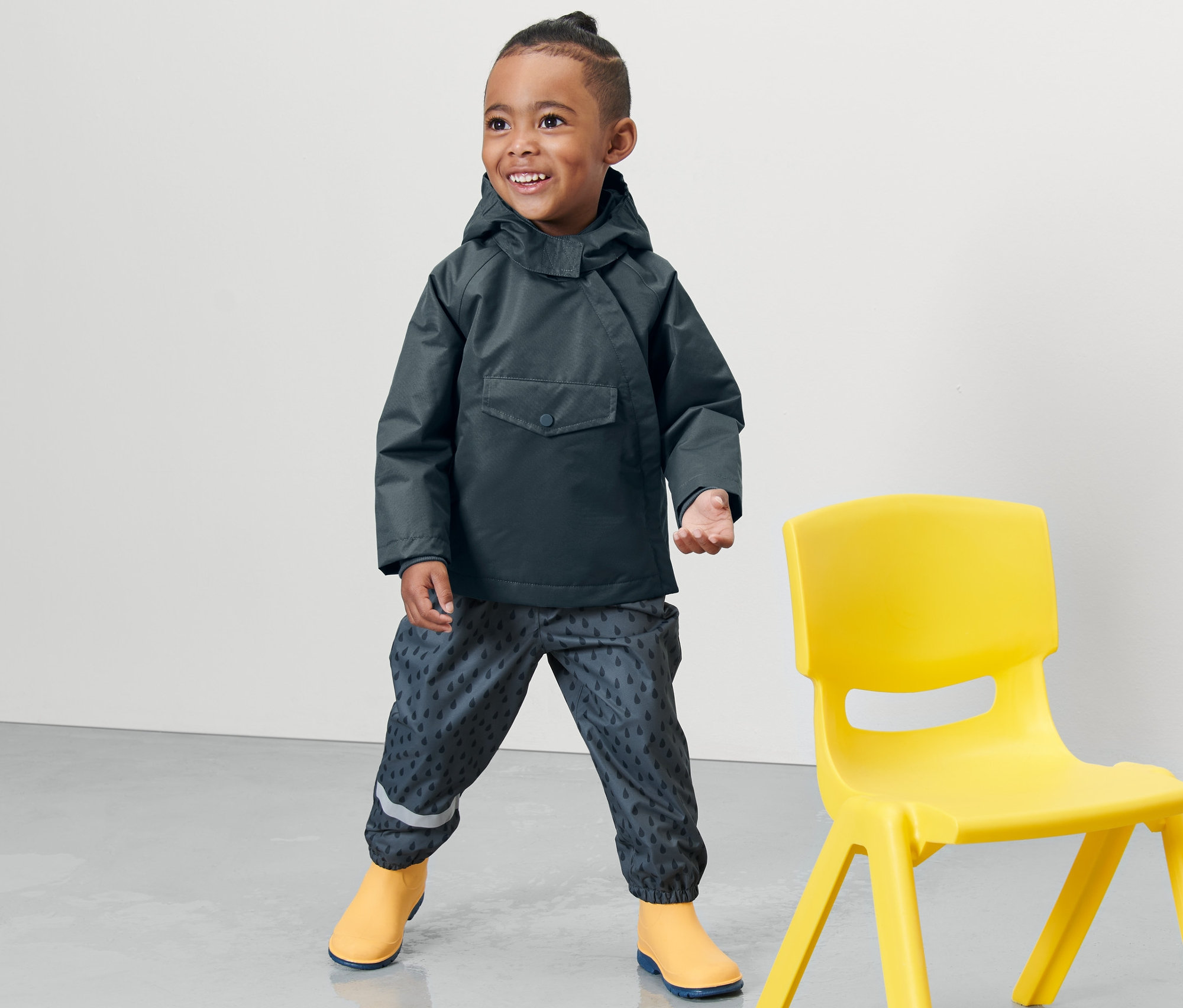Chłopiec stoi obok żółtego krzesła, ubrany w dziecięcą termiczną kurtkę przeciwdeszczową, łupkową niebieską.