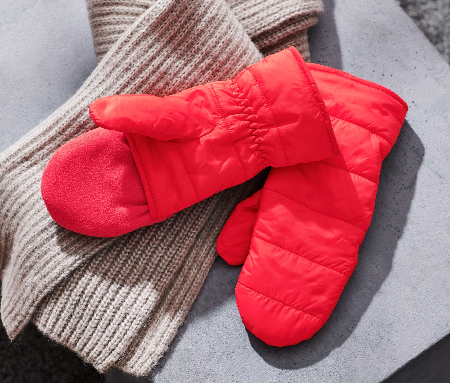 Na szarej powierzchni leżą czerwone rękawice-łapawice pikowane z jednym palcem i beżowy szalik z dzianiny.