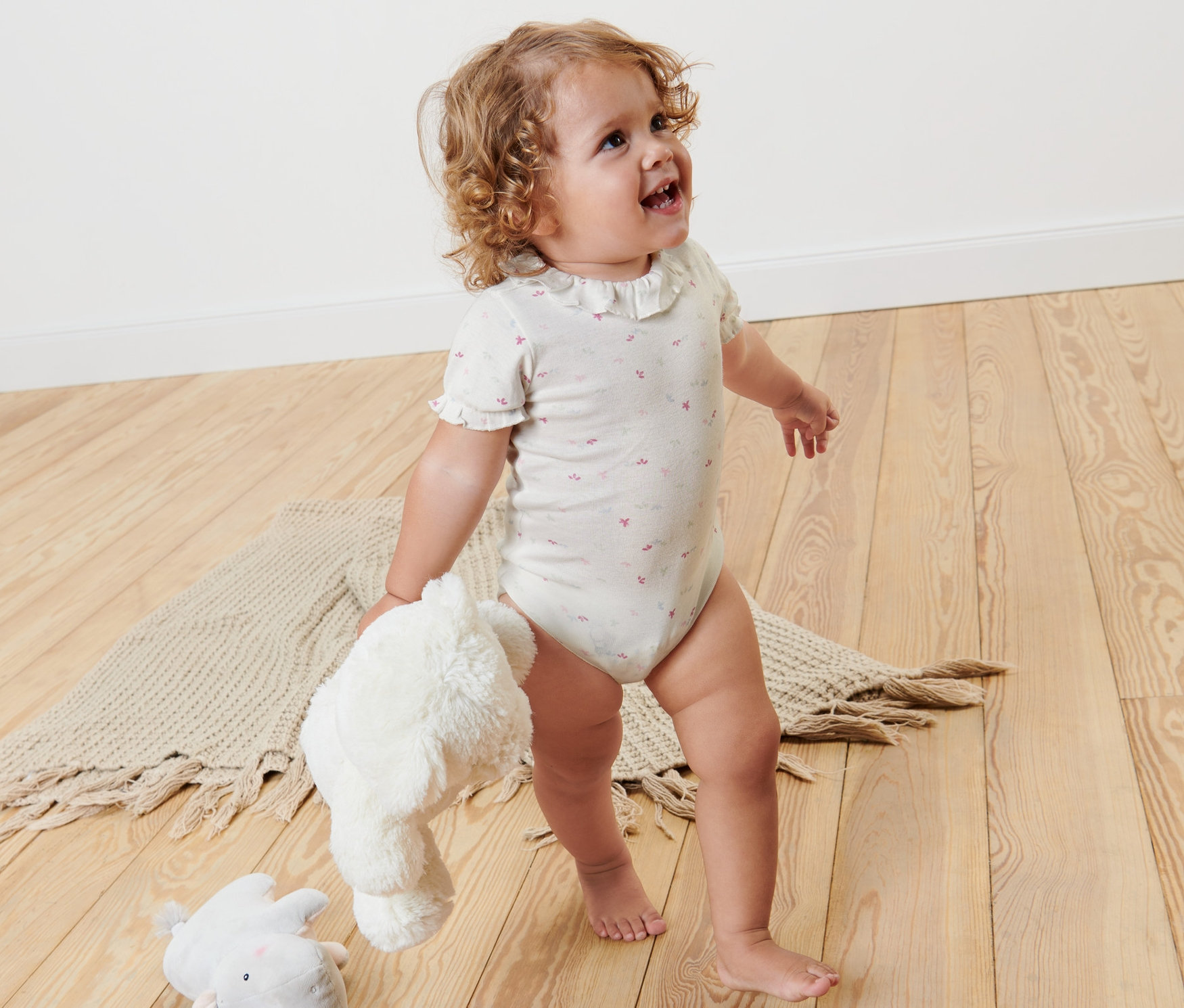 Małe dziecko stoi na drewnianej podłodze i trzyma pluszowe zabawki. Ubrane w body niemowlęce, 2 sztuki.