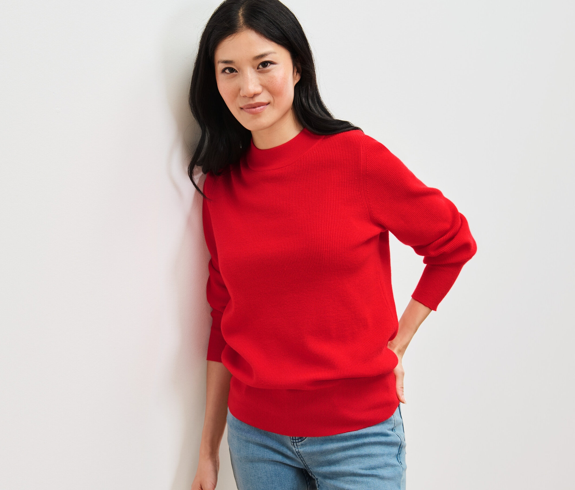 Kobieta opiera się o ścianę, ubrana w czerwony, cienki sweter i dżinsy.