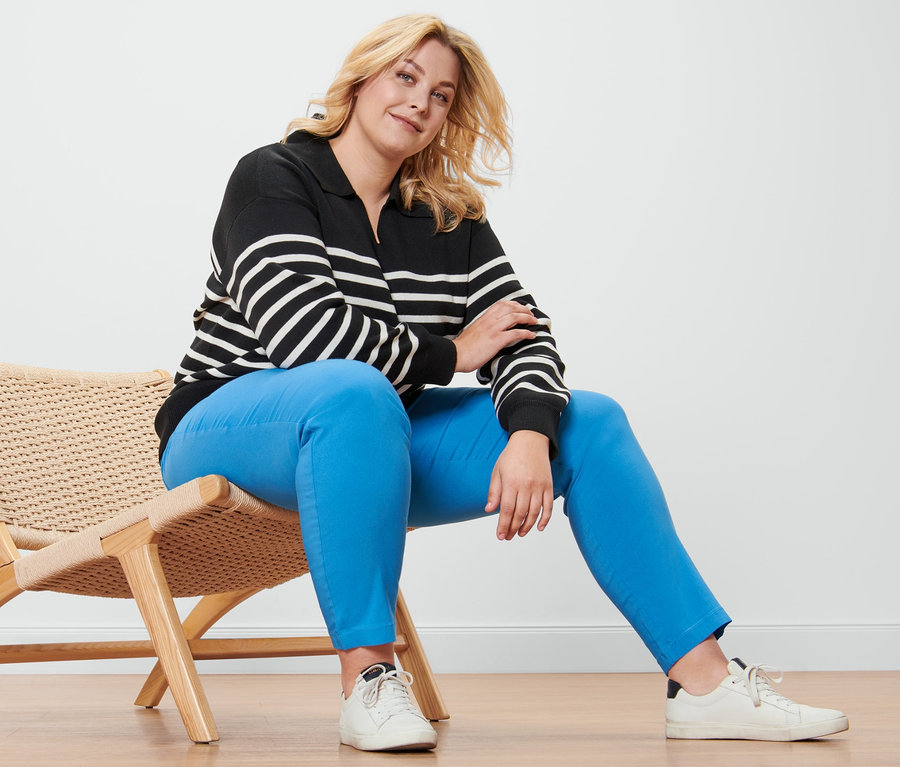 Blondwłosa kobieta siedzi na wiklinowym krześle i ma na sobie sweter z kołnierzem, spodnie z bengaliny i skórzane buty typu sneaker.