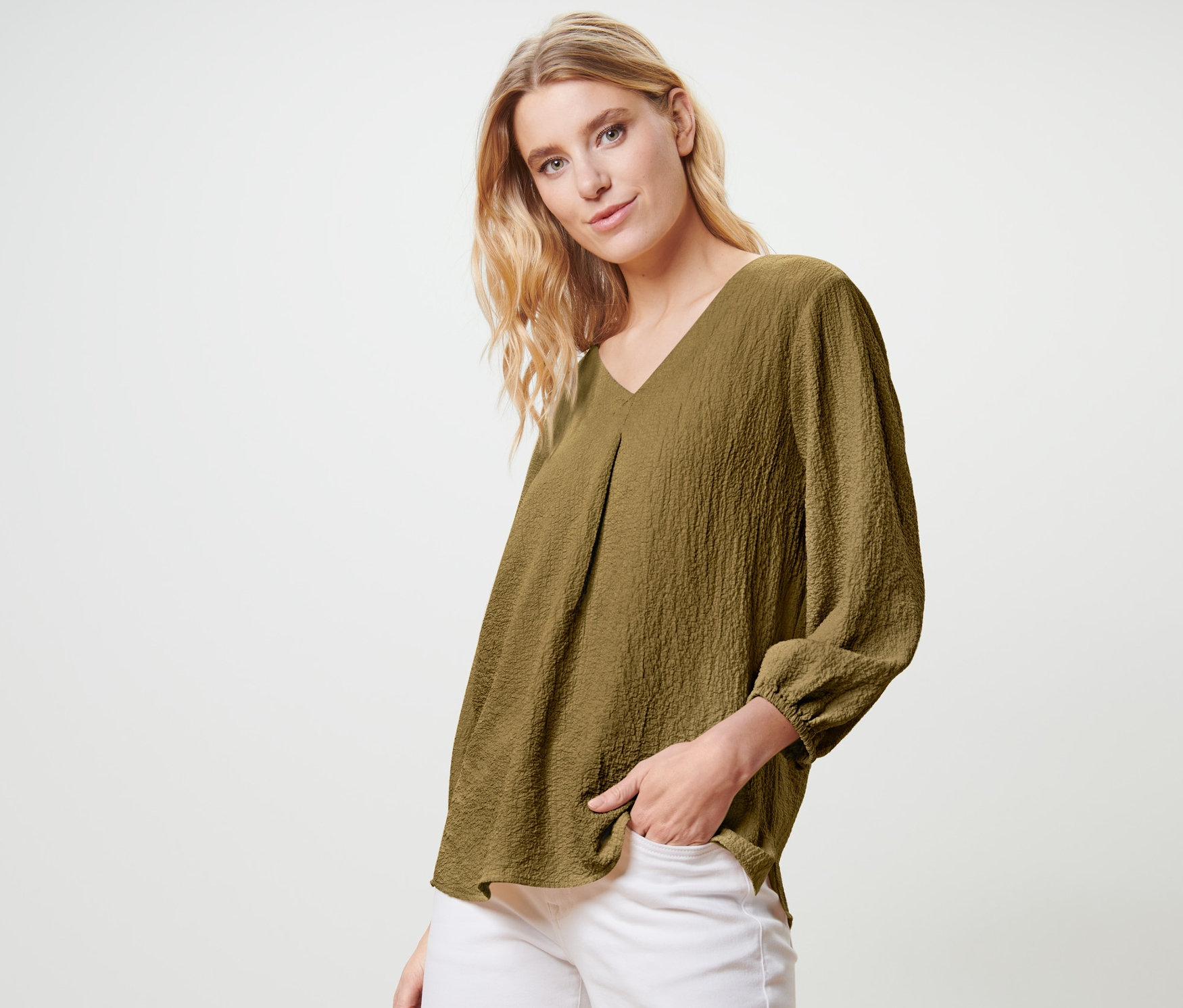 Blondynka pozuje w oliwkowozielonej bluzce-tunice i białych spodniach.