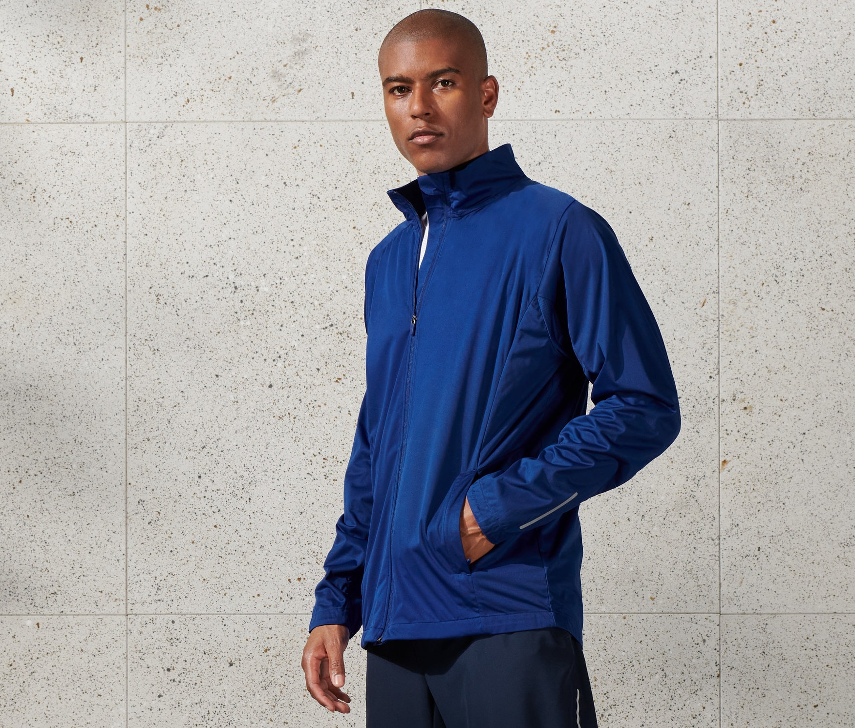 Mężczyzna ma na sobie niebieską, przeciwwiatrową kurtkę funkcyjną, szorty i białe tenisówki.