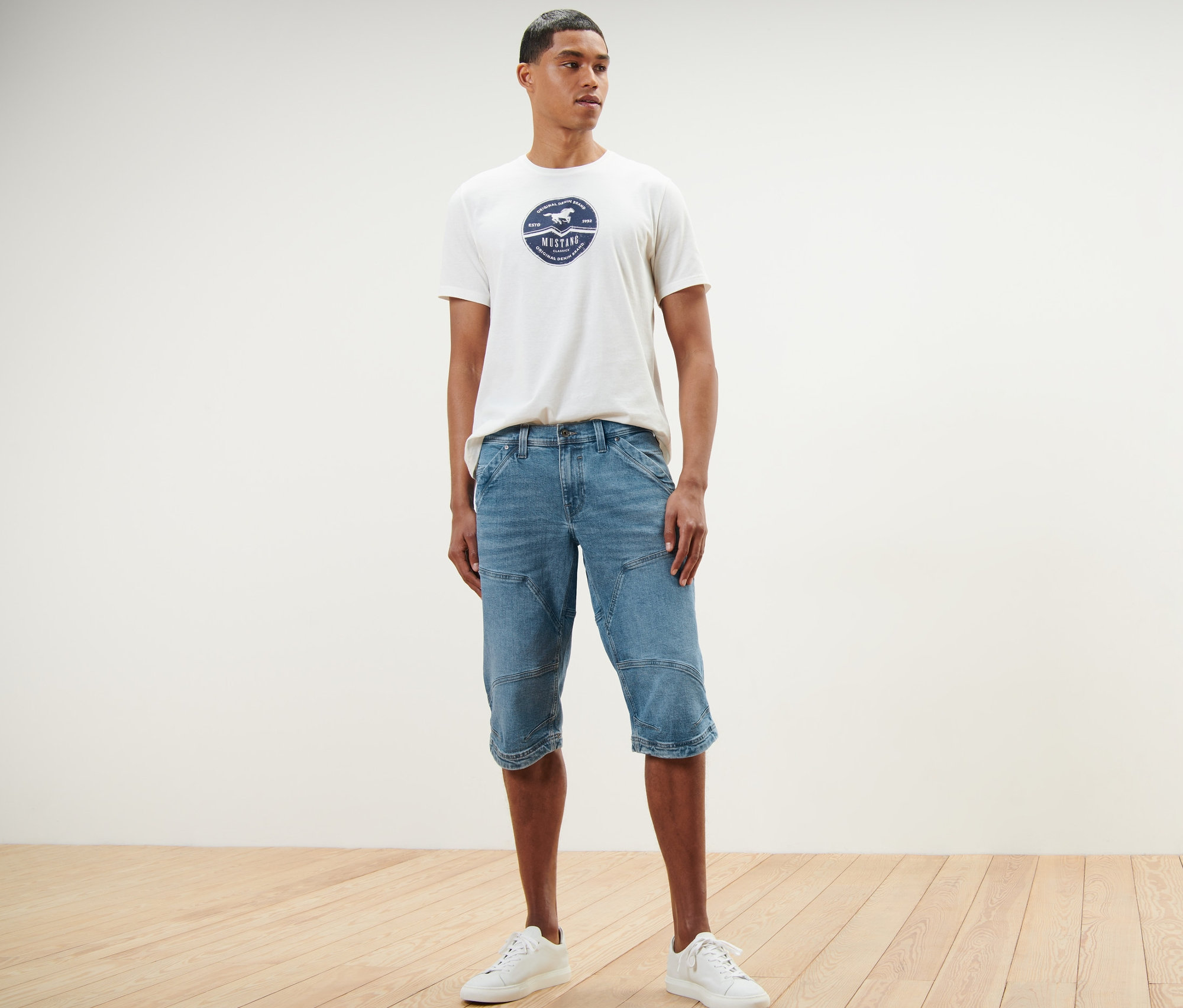 Mężczyzna stoi i ma na sobie dżinsowe szorty bermudy „Mustang” oraz T-shirt.