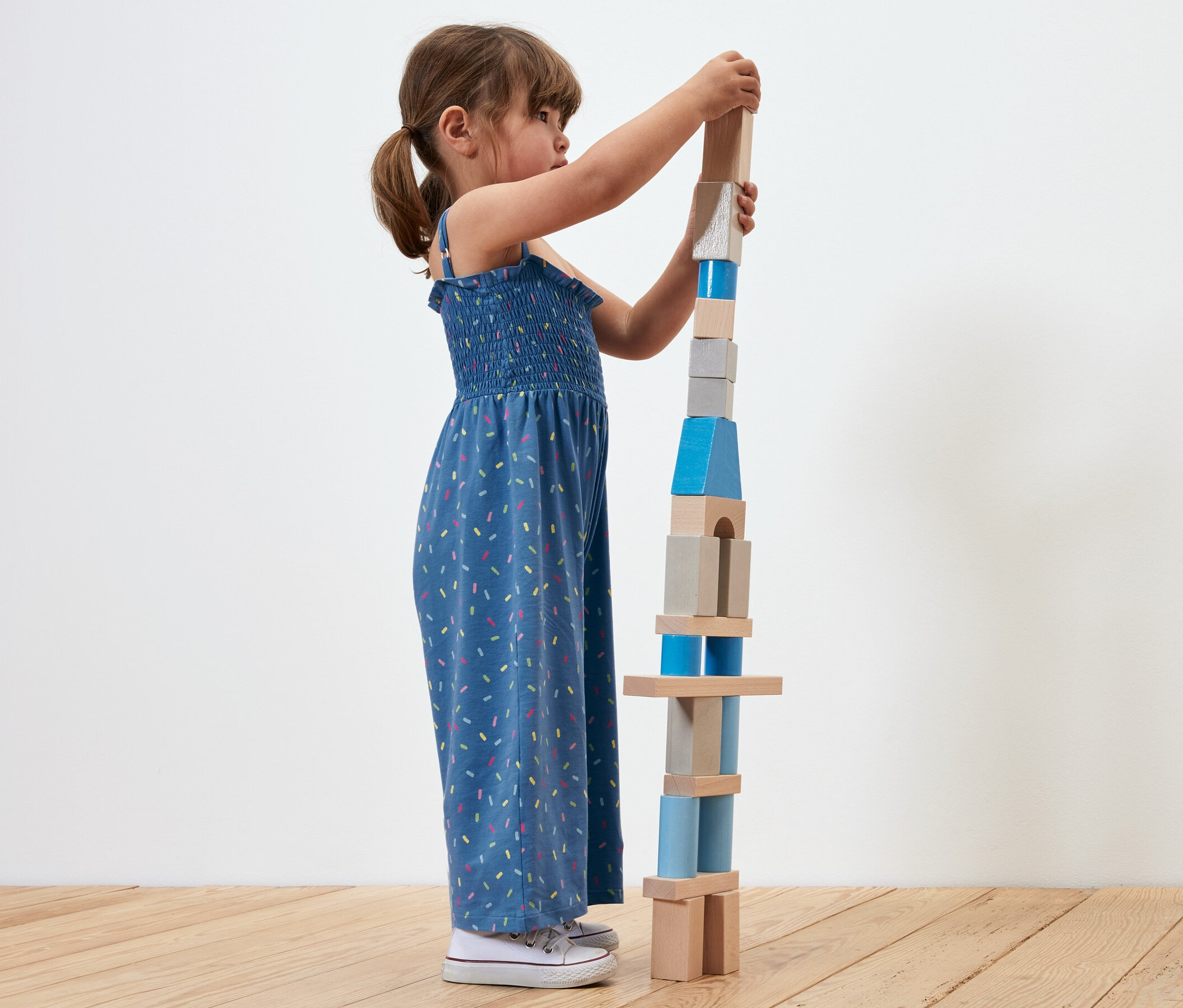 Dziewczynka buduje wieżę z drewnianych klocków. Ma na sobie kombinezon o długości 7/8.