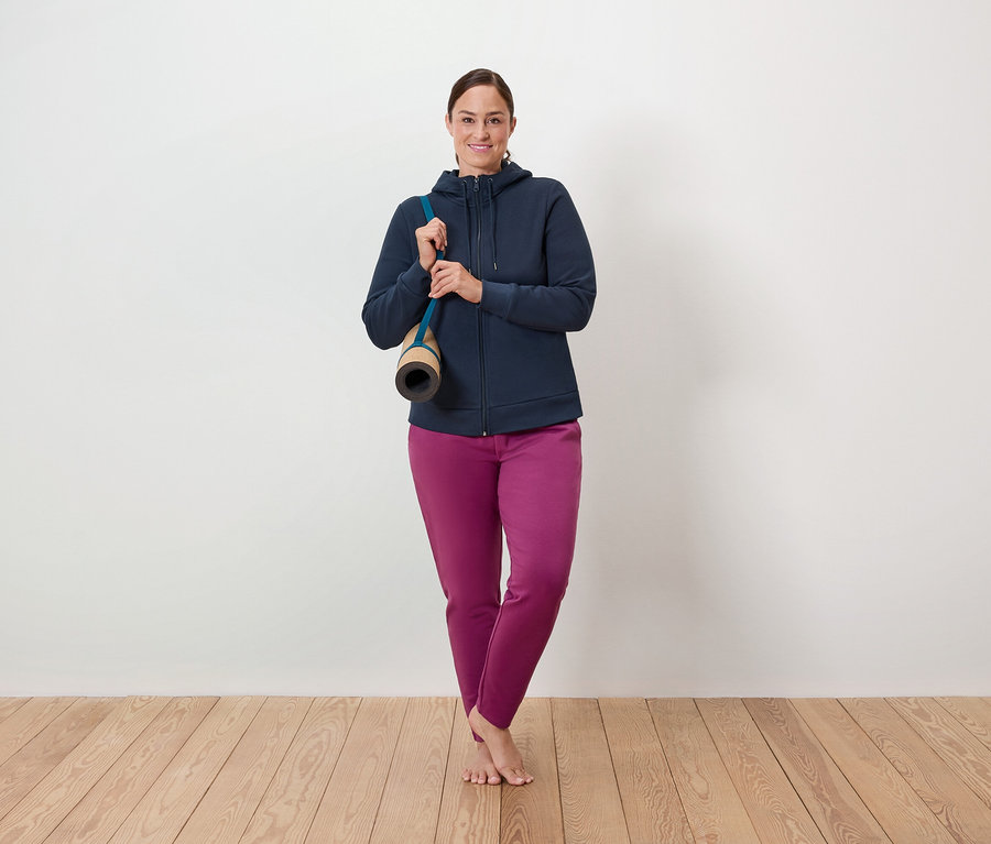 Kobieta stoi na drewnianej podłodze, z matą do ćwiczeń przewieszoną przez ramię, ubrana w niebieską bluzę z zamkiem błyskawicznym i fioletowe spodnie.