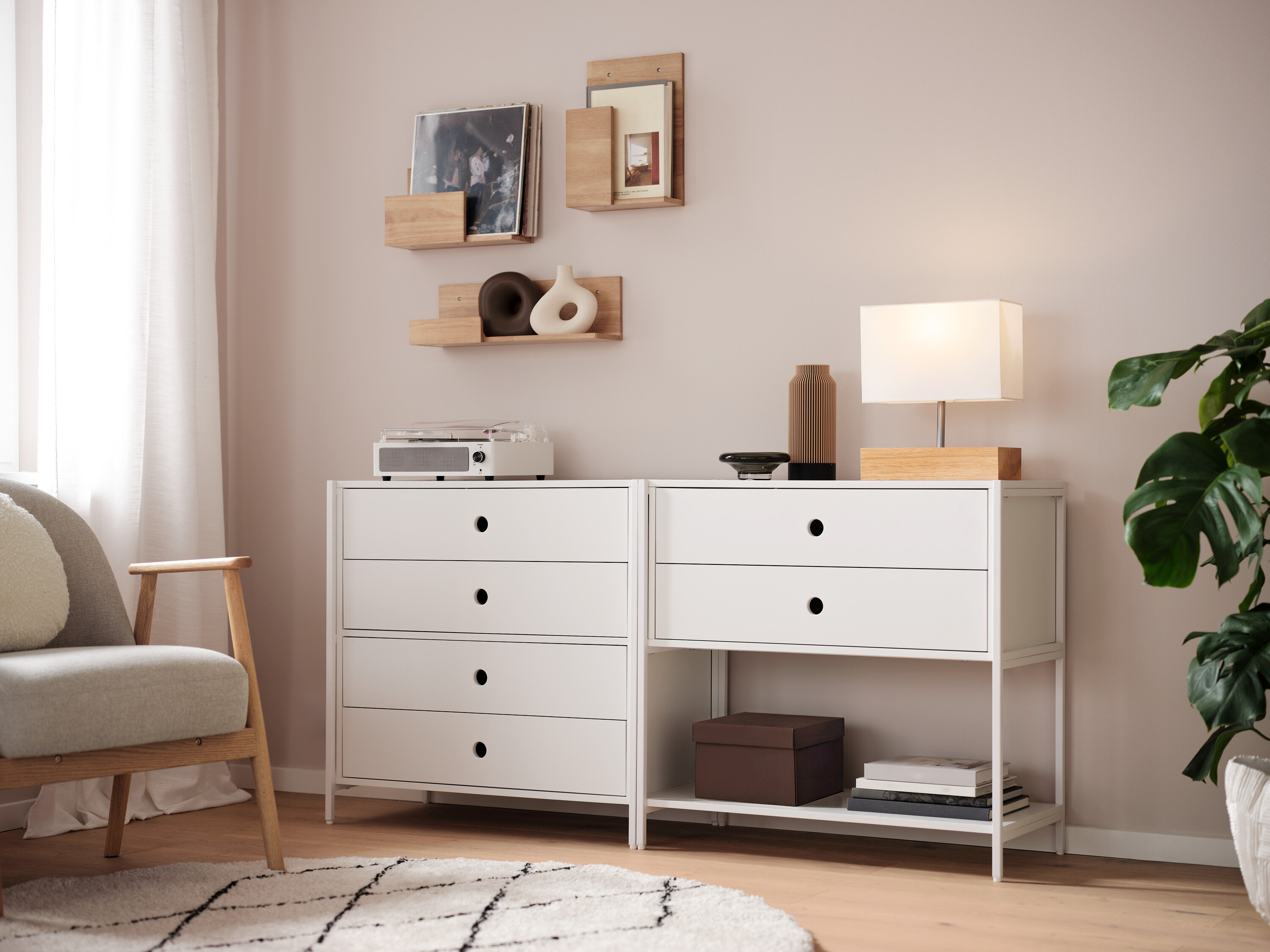 Dwie białe komody stoją obok siebie, na nich lampa, wazon i radio. Na ścianie wiszą półki.