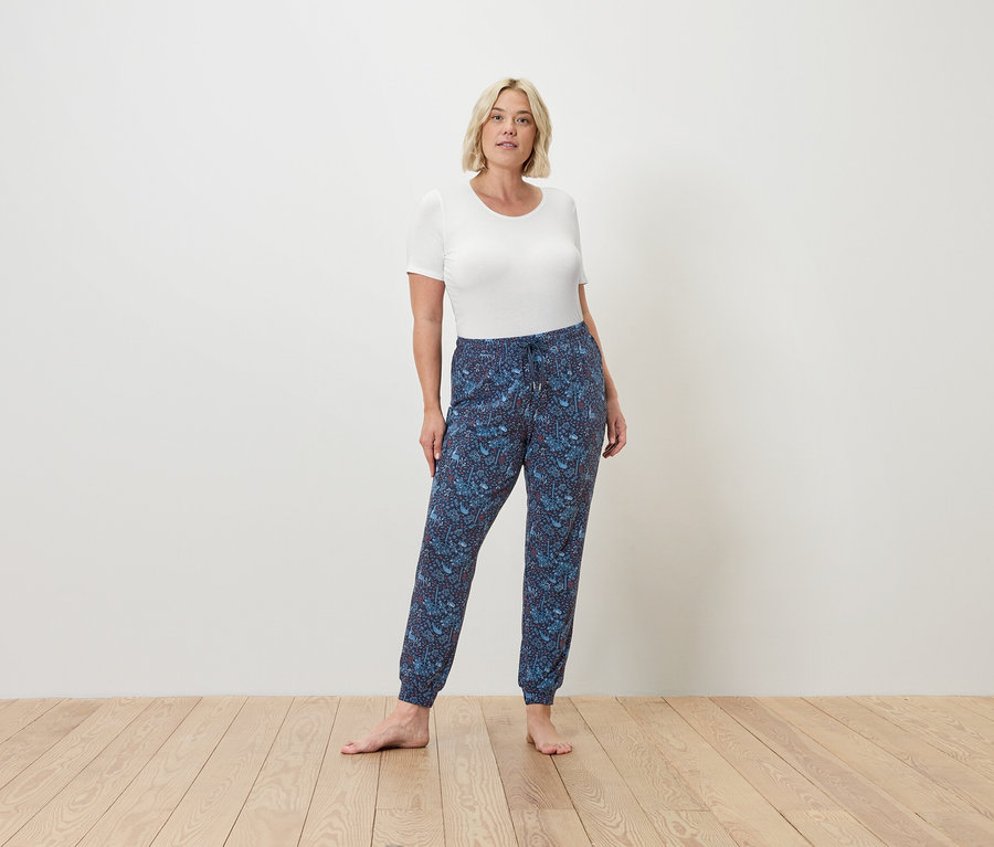 Kobieta stoi boso na drewnianej podłodze, ubrana w biały t-shirt i niebieskie spodnie typu jogger.