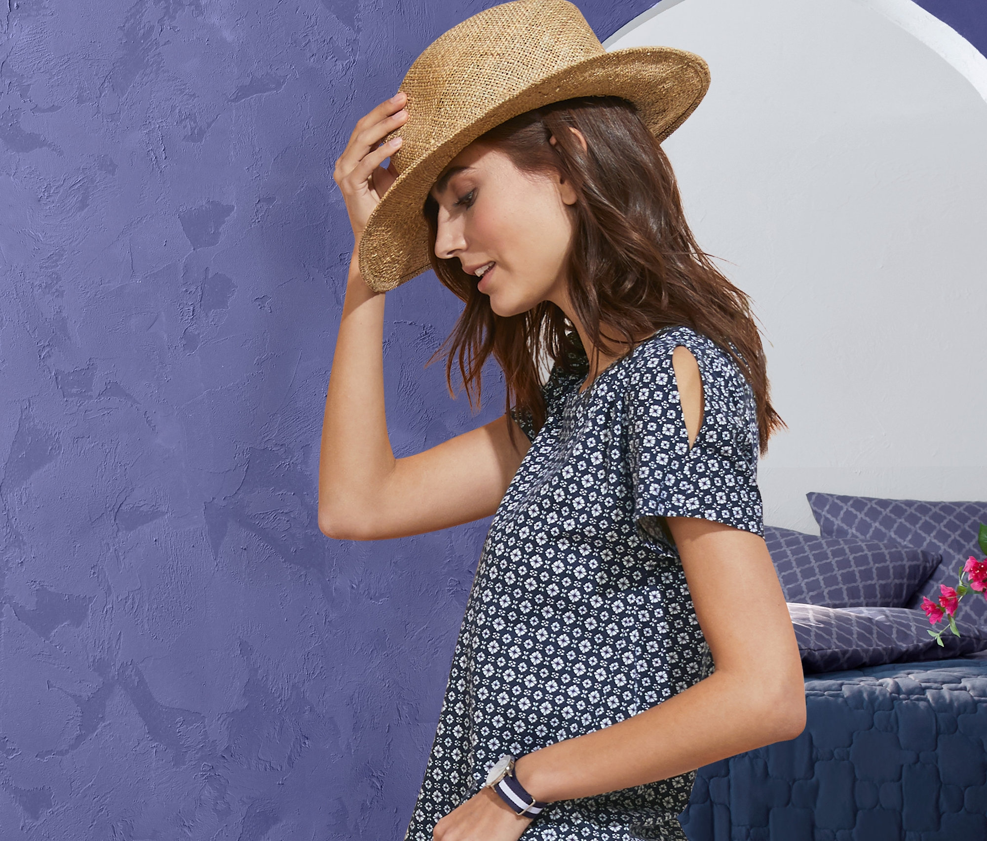 Kobieta trzyma kapelusz słomkowy, ma na sobie koszulkę z dekoracyjnym wzorem i elastyczne spodnie dżinsowe 7/8. W tle widać pościel, narzutę i pufę.