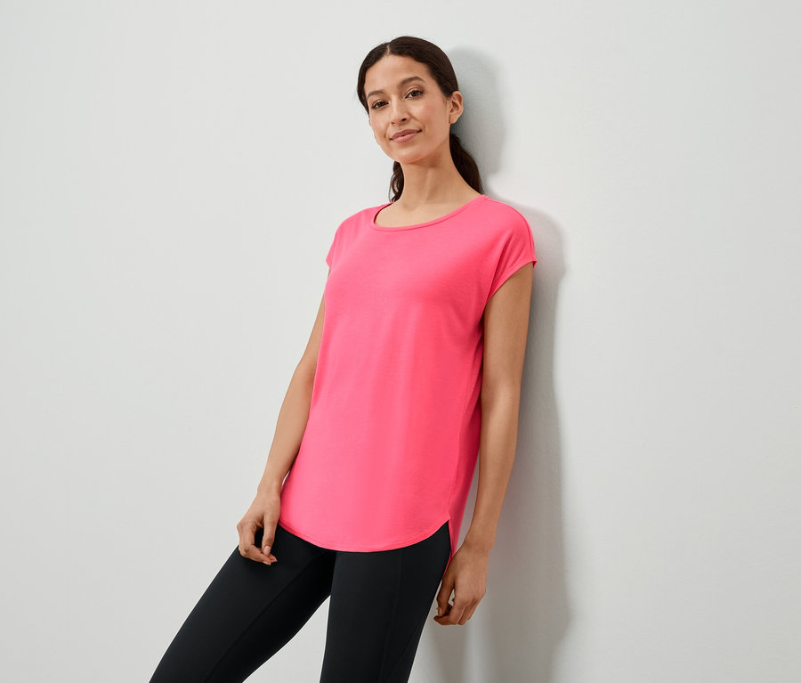 Kobieta opiera się o ścianę w różowej koszulce i czarnych legginsach.