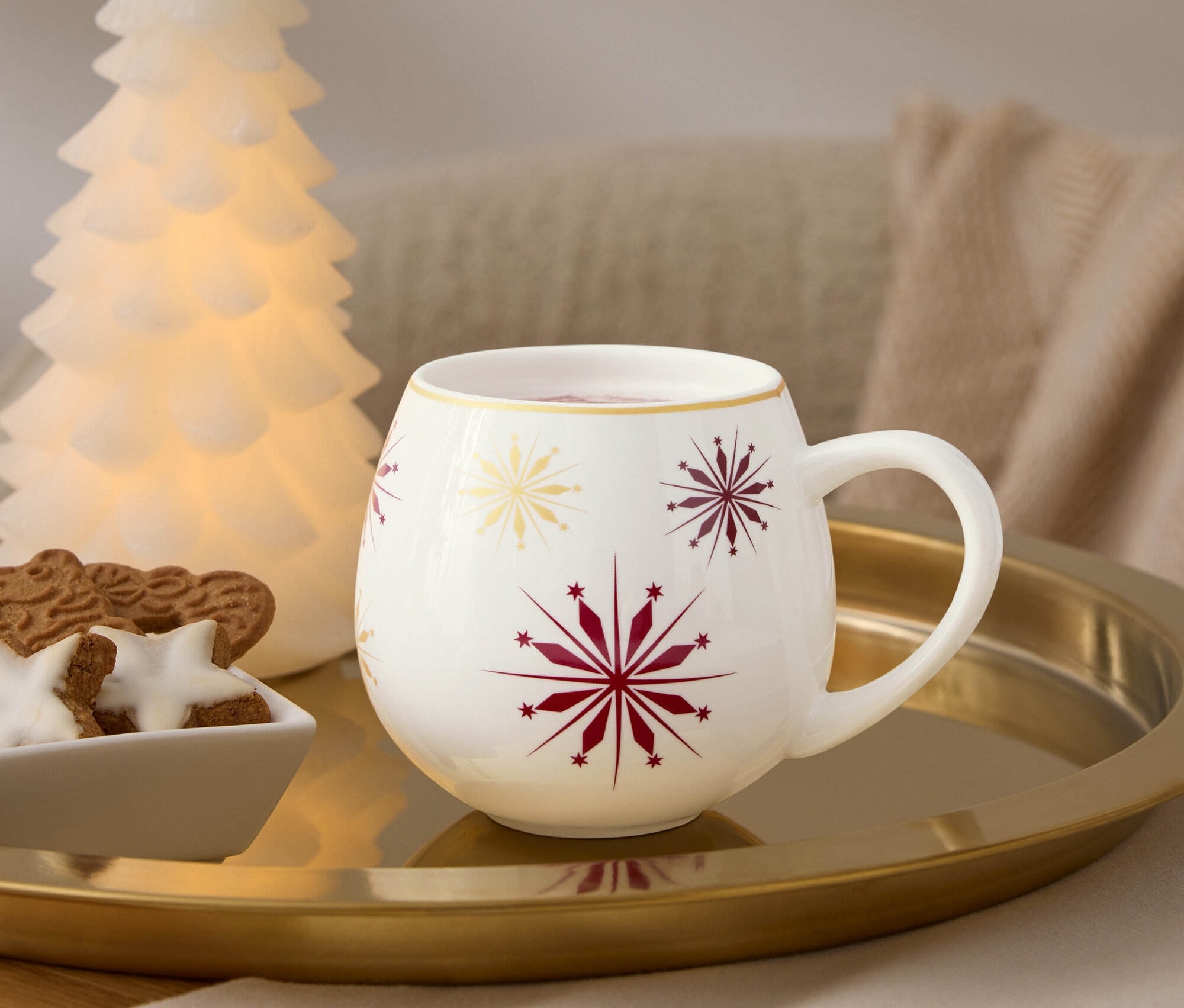 Na złotej tacy stoi biały kubek z czerwonymi i złotymi płatkami śniegu, obok niego miska z piernikami i świecąca choinka.
