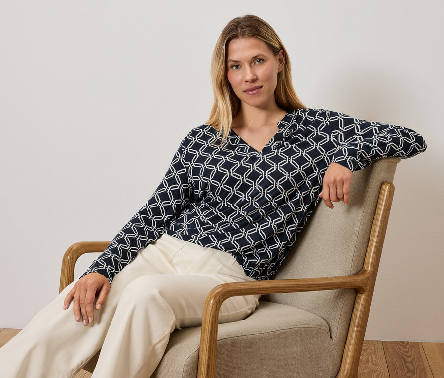 Kobieta siedzi w fotelu, ubrana w niebiesko-białą bluzkę i kremowe spodnie.