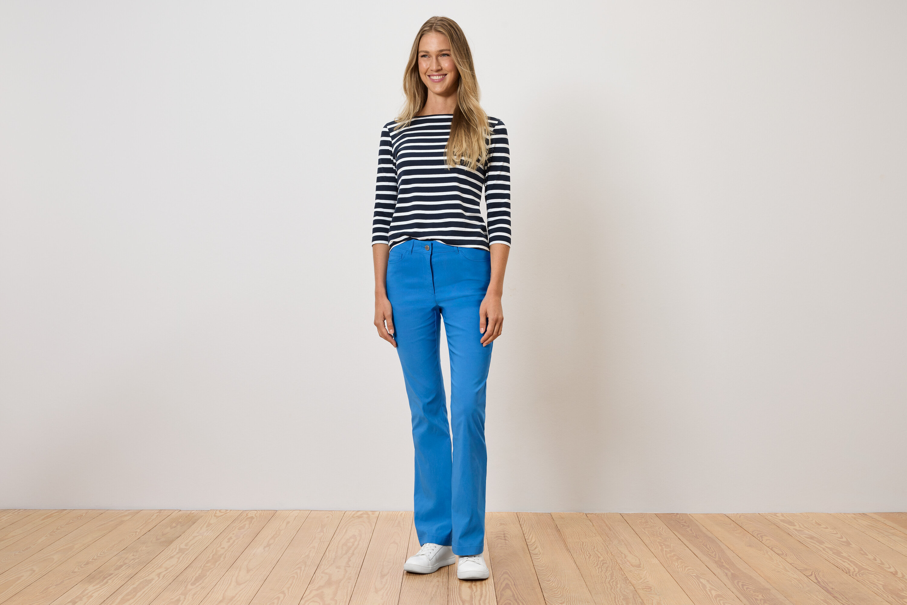 Kobieta stoi, ubrana w niebieskie spodnie, koszulkę w paski i białe buty.