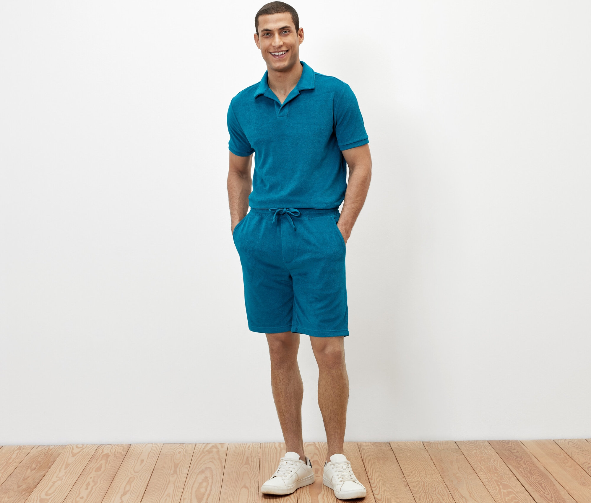 Mężczyzna stoi z rękami w kieszeniach szortów z tkaniny frotte w kolorze petrol.