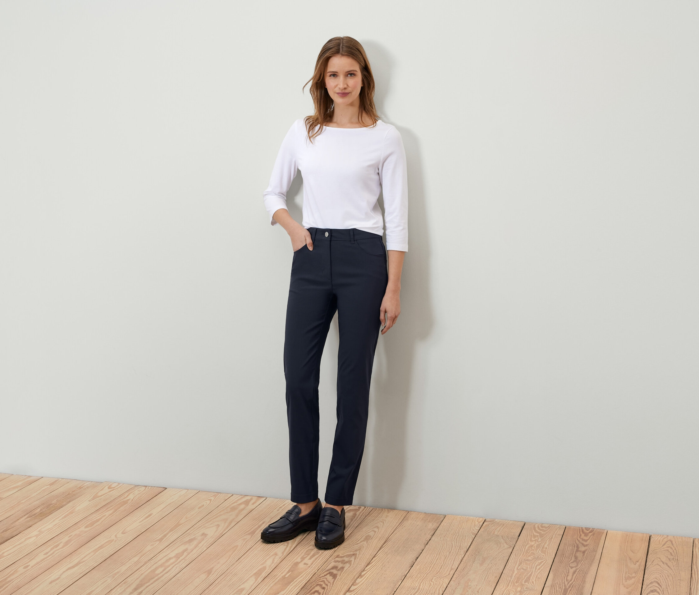 Kobieta stoi i pozuje w białej koszulce z rękawami trzy czwarte i granatowych spodniach.
