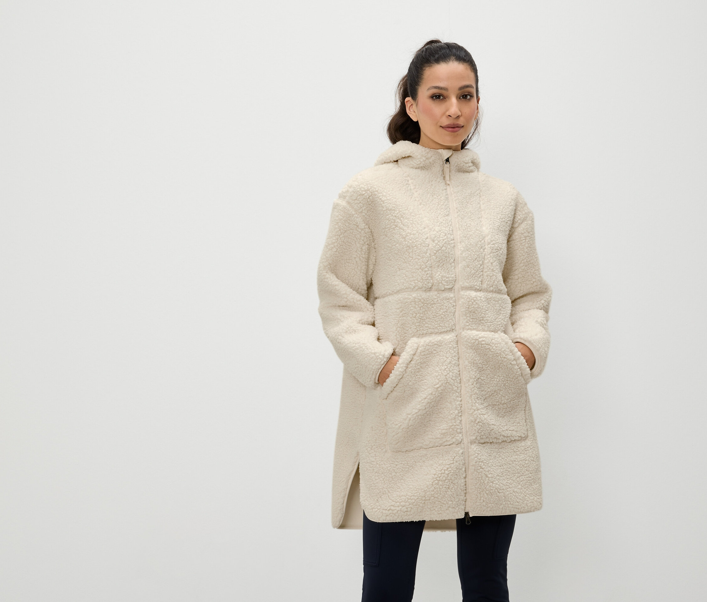 Kobieta stoi z rękami w kieszeniach kremowego płaszcza z kapturem zapinanego na zamek.