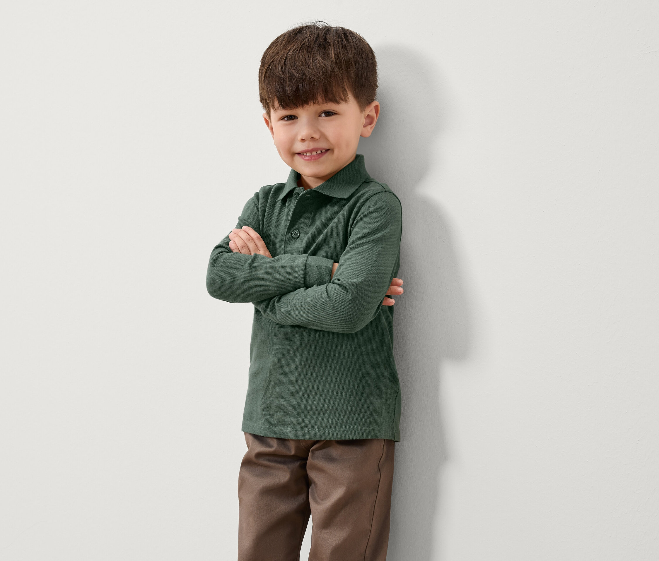 Chłopiec stoi pod ścianą ze skrzyżowanymi ramionami. Ma na sobie zieloną koszulkę polo z długim rękawem i brązowe spodnie.