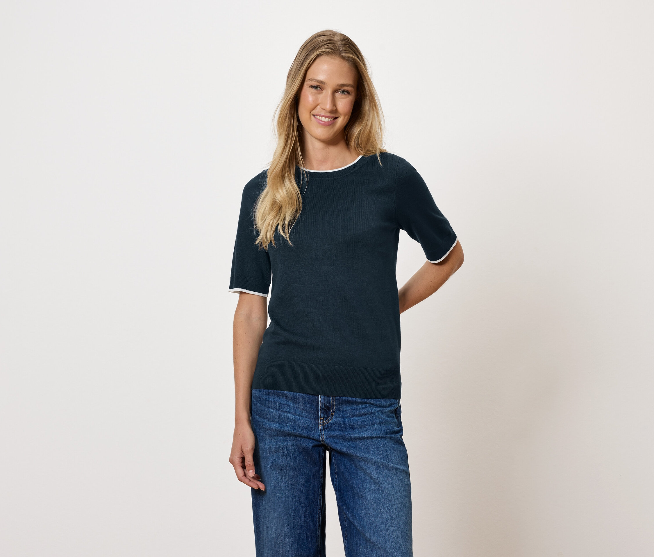 Kobieta z blond włosami ma na sobie granatową bluzkę z krótkim rękawem i dżinsy.