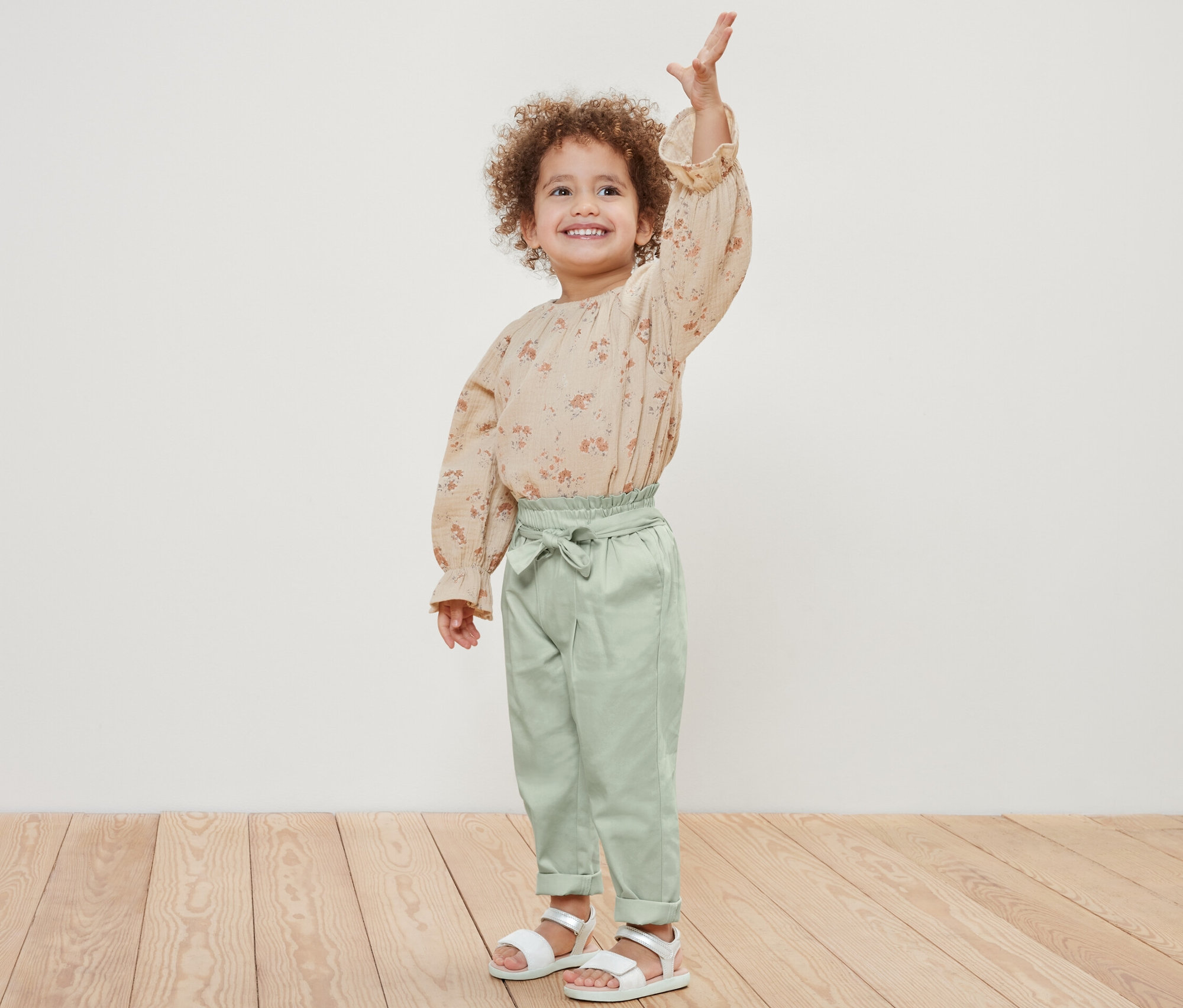 Uśmiechnięta dziewczynka pozuje i macha. Ma na sobie kwiecistą bluzkę i Spodnie na gumce.