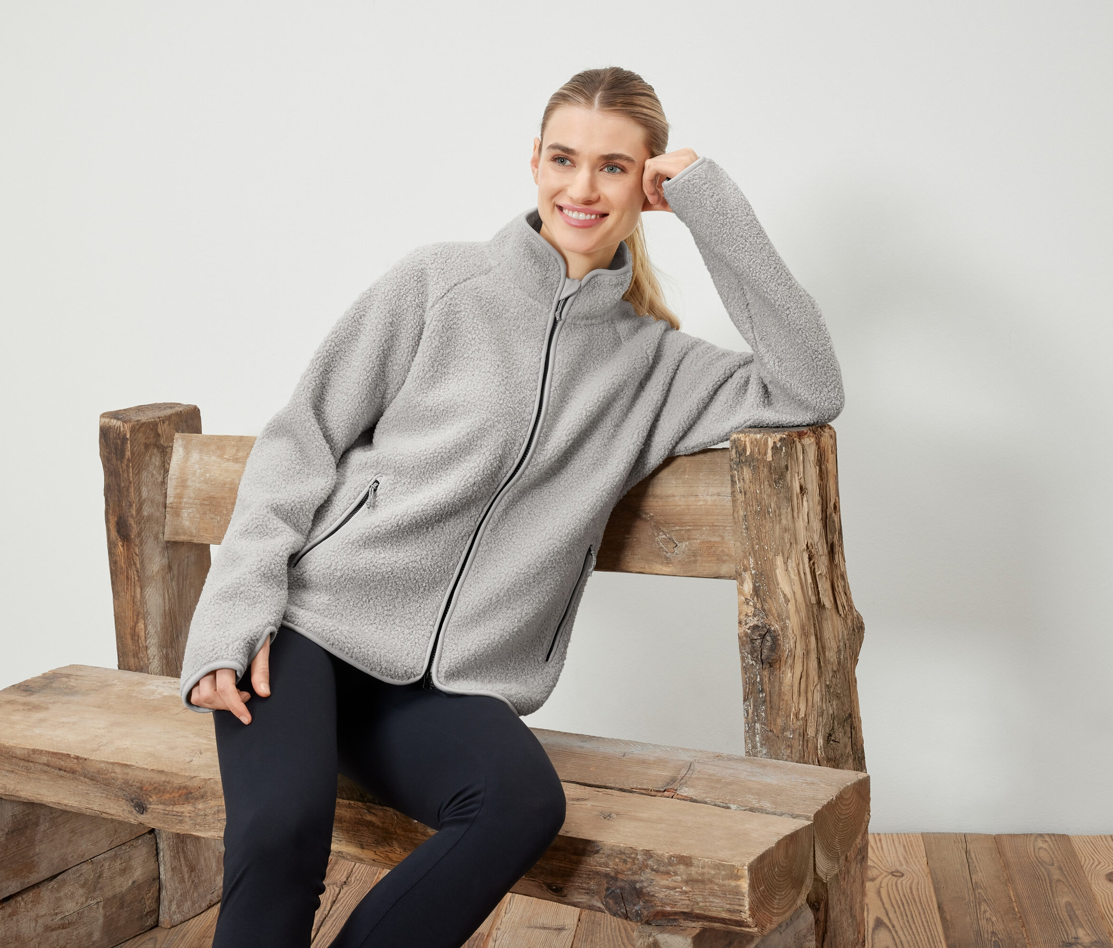 Kobieta w szarej bluzie polarowej pozuje na drewnianej ławce.