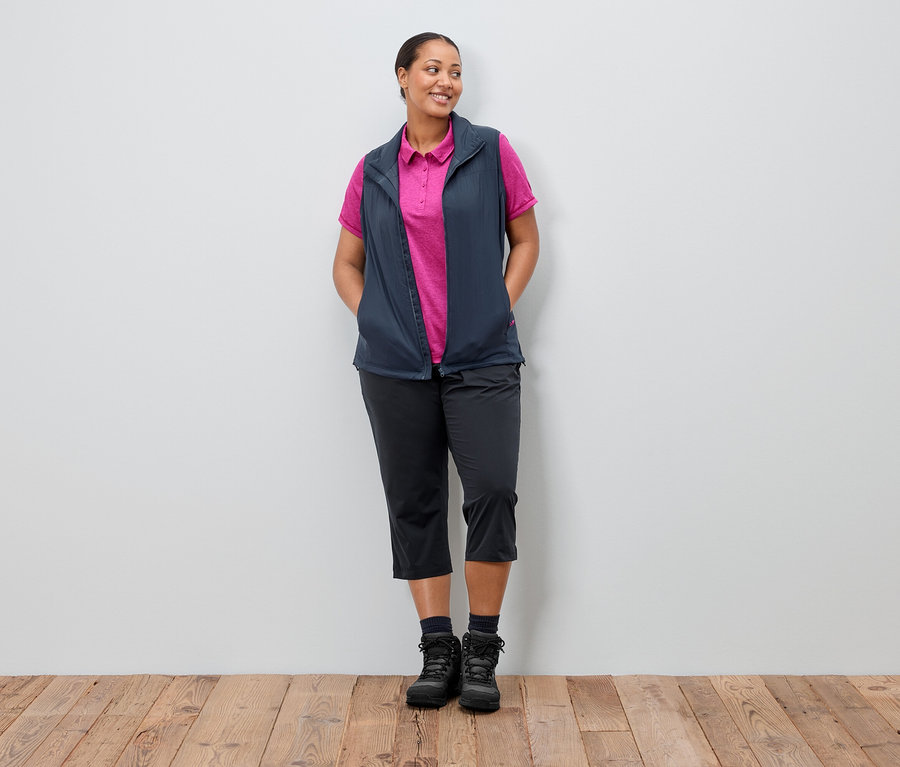 Kobieta stoi z rękami w kieszeniach na tle ściany. Ma na sobie granatową kamizelkę nałożoną na różową koszulkę polo, czarne spodnie i buty.