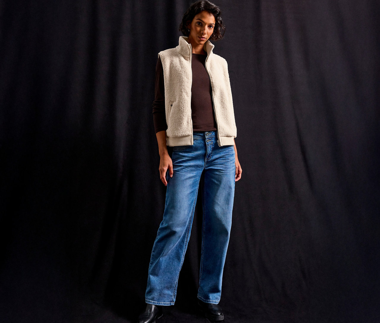 Kobieta ma na sobie pluszową kamizelkę Street One na brązową bluzkę z długim rękawem i dżinsy na czarnym tle.