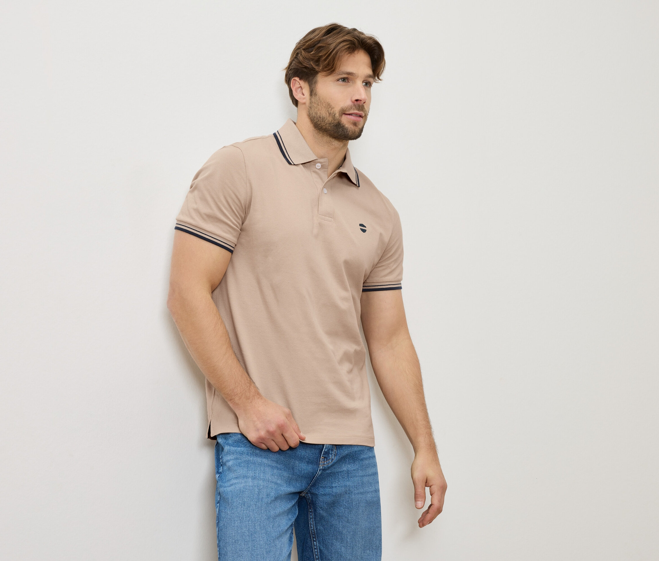 Mężczyzna w beżowej koszulce polo i dżinsach stoi przy ścianie.