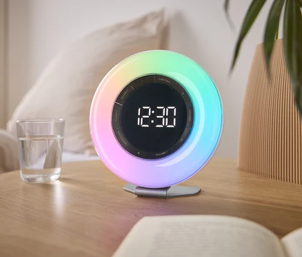 Na stoliku nocnym stoi kolorowy budzik świetlny wskazujący godzinę 12:30, obok szklanki wody i książki.