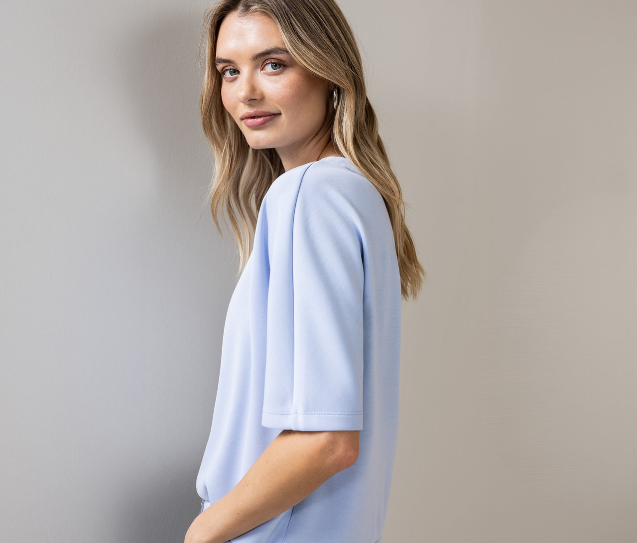 Kobieta spogląda przez ramię, ma na sobie błękitną koszulkę Street One z krótkimi rękawami i srebrne kolczyki.
