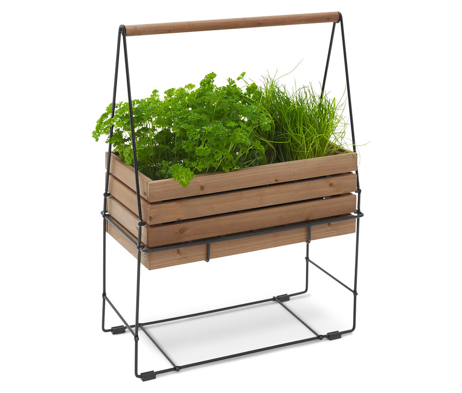 Drewniana skrzynka na zioła z pietruszką i szczypiorkiem, na czarnej metalowej ramie z drewnianym uchwytem.