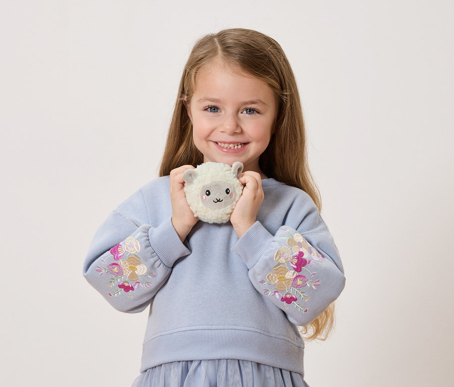 Uśmiechnięta dziewczynka trzyma przed sobą pluszową owieczkę. Ma na sobie niebieski sweter z kwiatowymi haftami na rękawach.